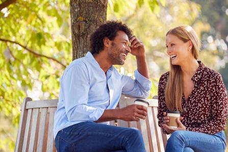 Two people smiling, talking, and drinking coffee on a park bench