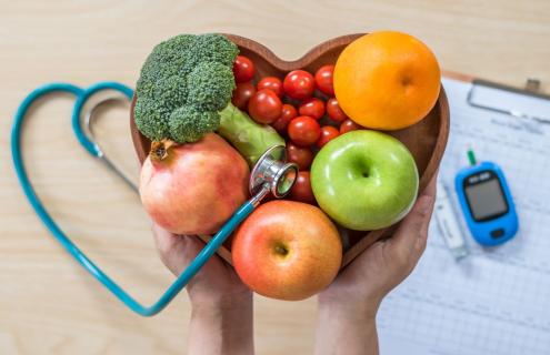 Image of hands holding a heart-shaped bowl filled with fruits and vegetables. Underneath is a stethoscope and glucose monitoring device.