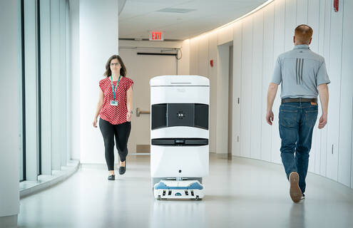 A TUG robot is seen traveling a hospital hallway with two people walking on either side of the robot.